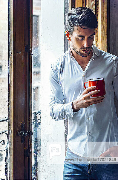 Nachdenklicher junger Mann mit Tasse am Fenster stehend