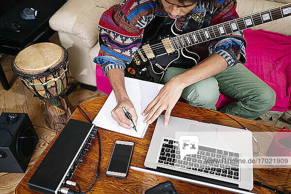 Junger Mann mit Gitarre schreibt Notizen auf einem Buch am Laptop im Wohnzimmer