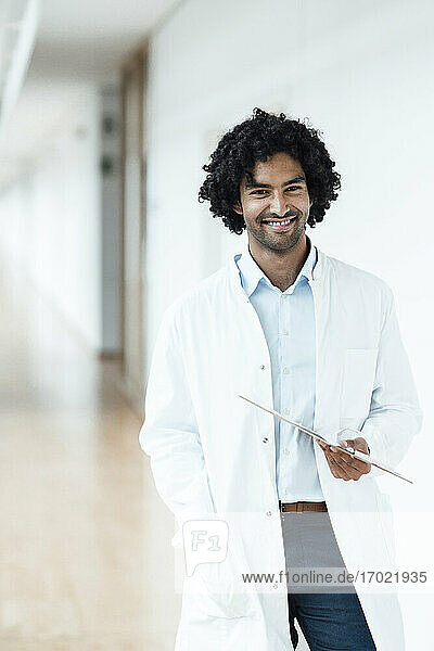 Smiling young male healthcare worker holding clipboard while standing at hospital corridor