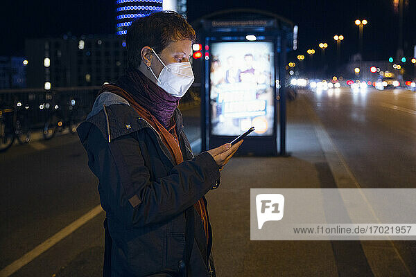 Frau mit Gesichtsmaske  die ein Mobiltelefon benutzt  während sie an einer Bushaltestelle in der Stadt steht