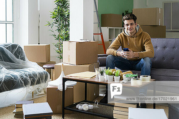 Lächelnder junger Mann mit Smartphone  der in einem Raum voller Kisten sitzt und umzieht