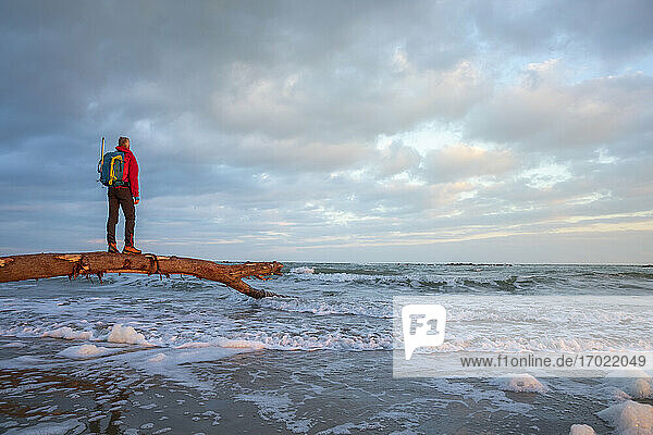 Male hiker standing on log over Mediterranean Sea against cloudy sky  Italy