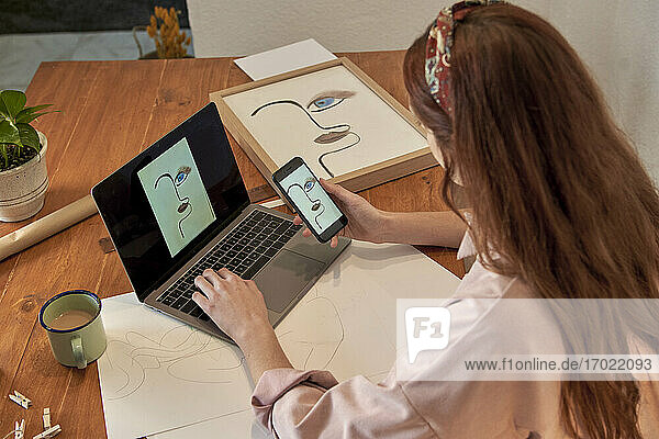 Künstlerin  die ein Foto von einer Zeichnung online über einen Laptop am Schreibtisch in ihrem Haus teilt