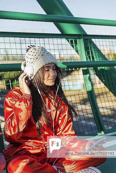 Woman wearing headphones and knit hat listening music while sitting on bridge