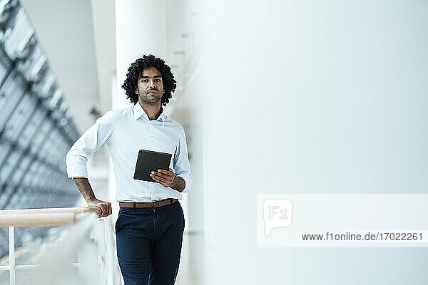 Selbstbewusster Geschäftsmann  der ein digitales Tablet in der Hand hält  während er an einem Geländer im Korridor eines Büros steht