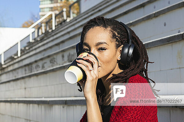 Junge Frau hört Musik und trinkt Kaffee aus einem wiederverwendbaren Becher an einem sonnigen Tag