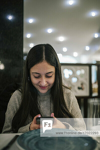 Lächelndes Teenager-Mädchen  das ein Mobiltelefon benutzt  während es im Restaurant sitzt