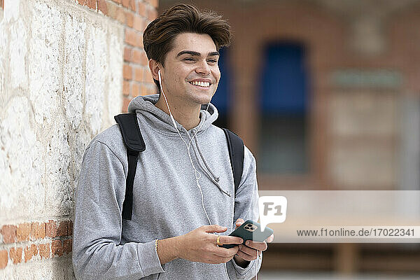 Cheerful handsome young man listening music while holding smart phone against wall