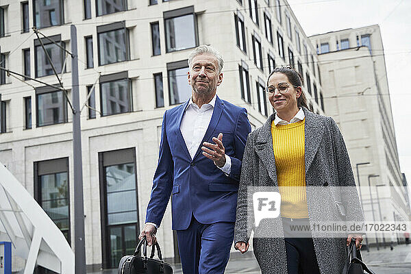 Lächelnde männliche und weibliche Geschäftskollegen unterhalten sich beim Spaziergang gegen ein Gebäude in der Stadt