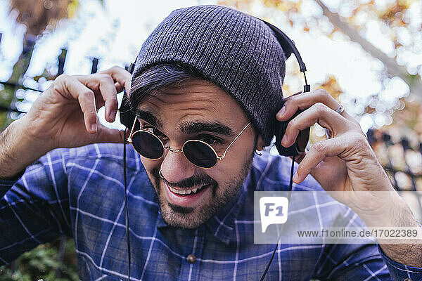 Lächelnder junger Mann mit Sonnenbrille und Kopfhörern in einem öffentlichen Park
