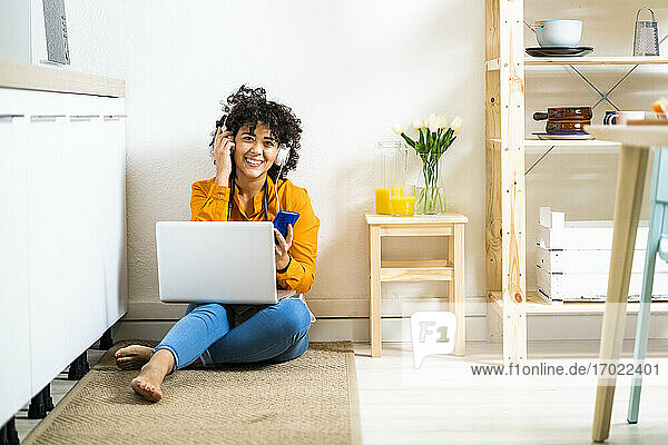 Junge Frau mit Kopfhörern  die ein Mobiltelefon benutzt  während sie zu Hause mit einem Laptop auf dem Boden sitzt