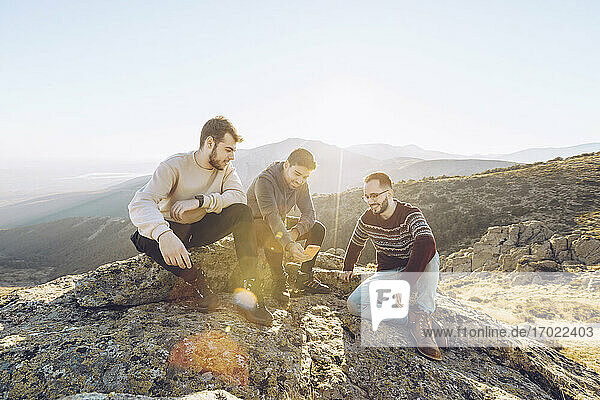 Male friends talking while sitting on mountain against clear sky during summer