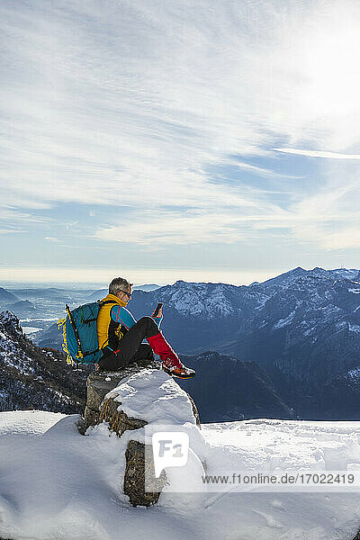 Älterer Mann mit Rucksack  der ein Mobiltelefon benutzt  während er auf einem verschneiten Berg sitzt  Orobic-Alpen  Lecco  Italien