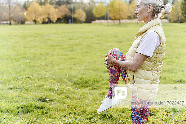Ältere Frau  die sich auf einem Bein stehend über Gras in einem öffentlichen Park streckt