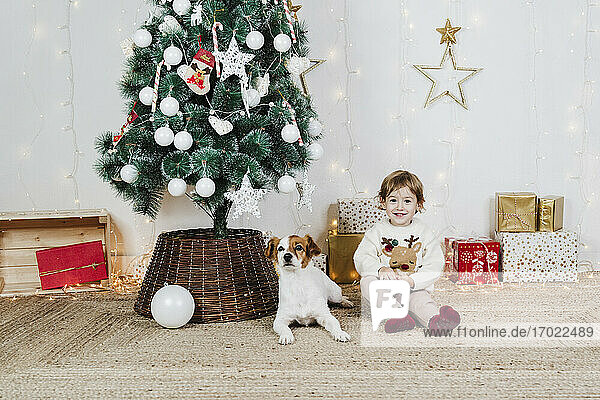 Smiling baby girl with puppy sitting by Christmas tree and gifts at home