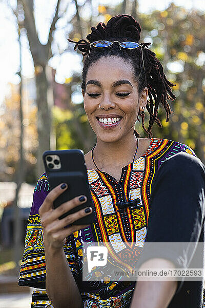 Glückliche modische junge Frau  die im Park sitzend ein Mobiltelefon benutzt