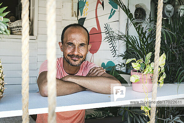 Lächelnder reifer Mann  der sich auf seiner Terrasse an ein Pflanzenregal lehnt