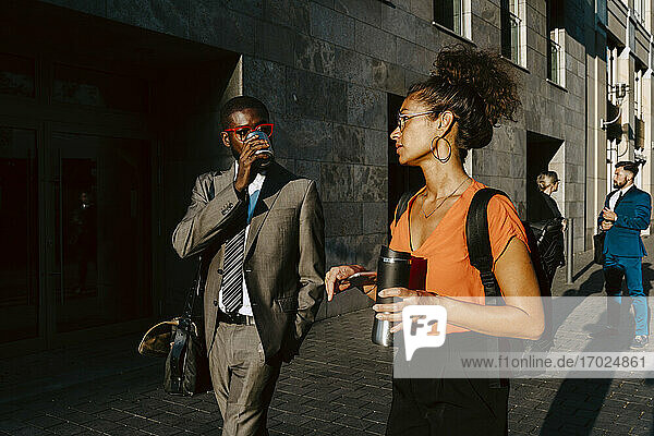 Männlicher Unternehmer trinkt Kaffee  während er mit einer weiblichen Kollegin auf einem Fußweg in der Stadt diskutiert