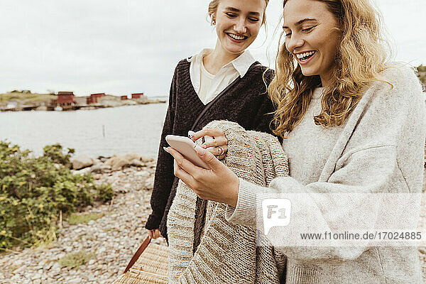Smiling female friends using smart phone against sea during vacation