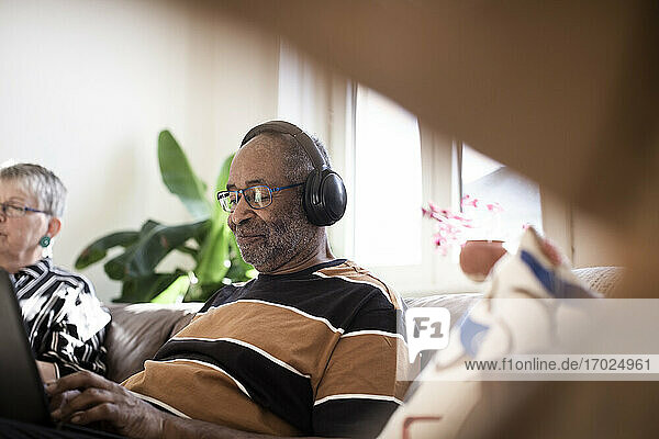Lächelnder älterer Mann  der Musik über Kopfhörer hört und neben einer Frau auf dem Sofa im Wohnzimmer sitzt