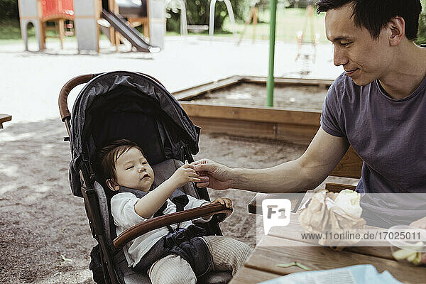 Father feeding baby son while sitting in public park