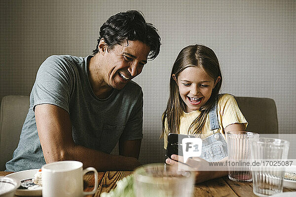 Smiling father and daughter using mobile phone at dining table