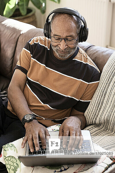 Älterer Mann hört Musik über Kopfhörer  während er einen Laptop im Wohnzimmer benutzt