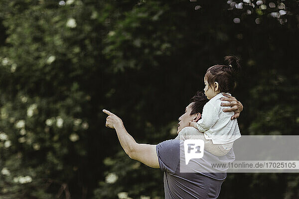 Vater zeigt auf ein männliches Kleinkind  während er es im Park über der Schulter trägt