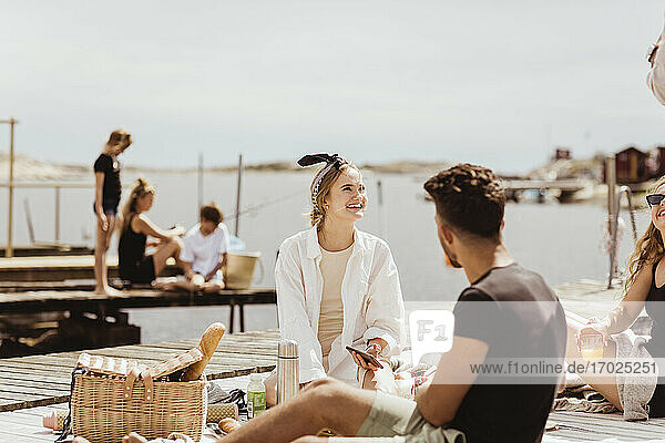 Smiling friends talking while sitting over jetty on sunny day during picnic