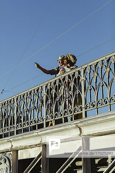 Älterer Mann  der auf eine Frau auf einer Brücke gegen den blauen Himmel zeigt