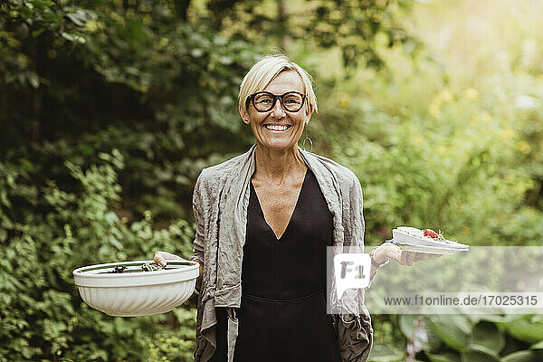 Lächelnde reife Frau hält Essen in Schüssel und Teller gegen Pflanzen im Vorgarten