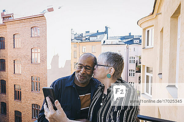 Frau küsst Mann während der Aufnahme von Selfie auf Smartphone in Balkon