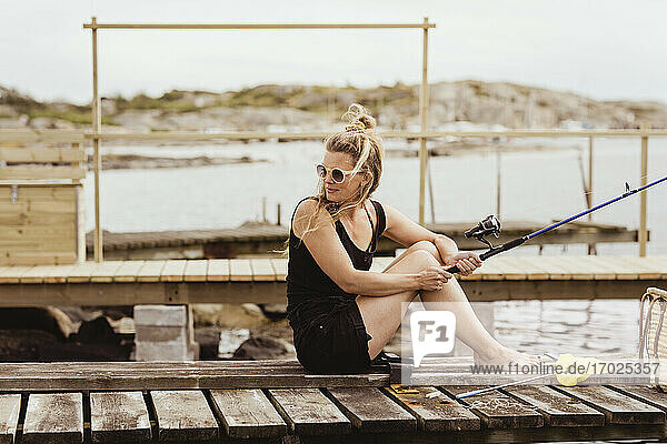 Mature woman with fishing rod looking over shoulder while sitting on jetty at harbor