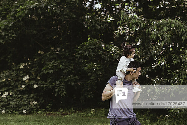 Vater schaut weg  während er seinen Sohn über die Schulter gegen Pflanzen im Park trägt