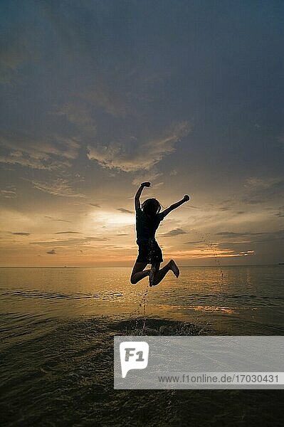 Eine junge Frau springt vor Freude und feiert am Strand bei Sonnenuntergang.