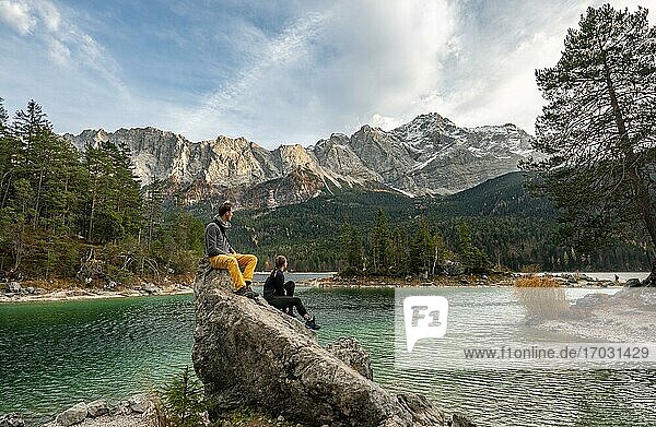 Young woman and young man sitting on a rock at the shore  view into the distance  Eibsee lake in front of Zugspitzmassiv with Zugspitze  Wetterstein Mountains  near Grainau  Upper Bavaria  Bavaria  Germany  Europe