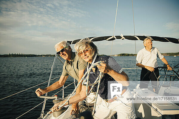 Senior man turning handle on boat while female friend with rope standing by against sky