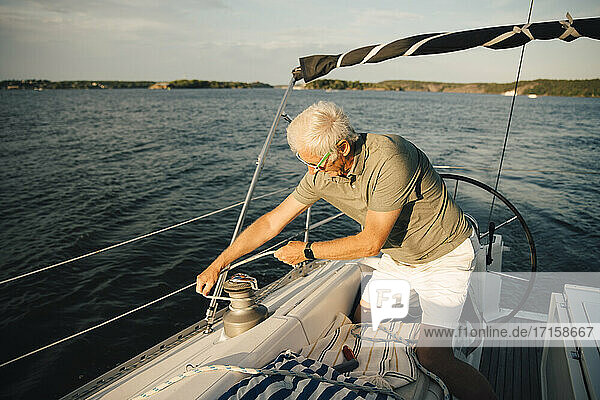 Senior Mann bindet Seil auf Klampe  während kniend in Boot während sonnigen Tag