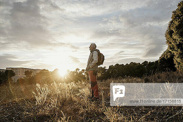 Senior male hiker with hands in pockets standing against cloudy sky at countryside during sunset