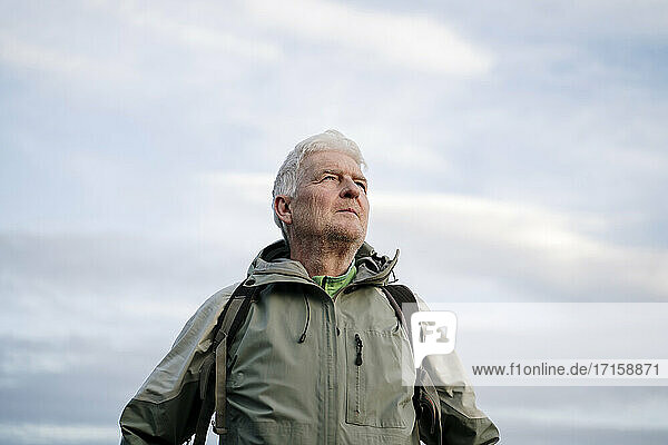 Senior male hiker looking away while standing against cloudy sky during weekend