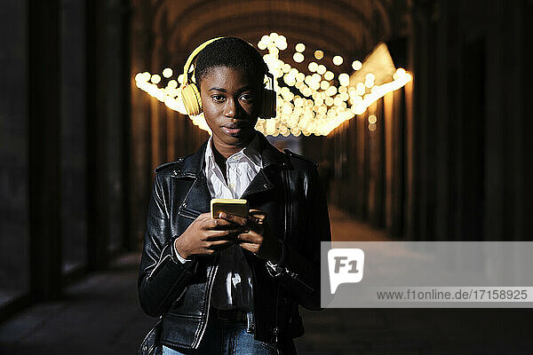 Young woman with headphones and mobile phone staring while standing at corridor