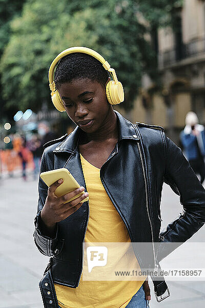 Young woman wearing headphones using smart phone while standing with hand on hip in city