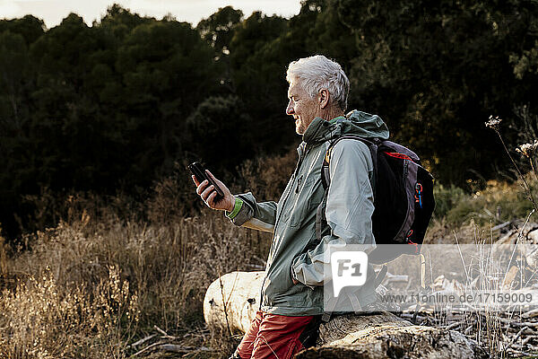 Senior man using mobile phone on wooden log in field during sunset