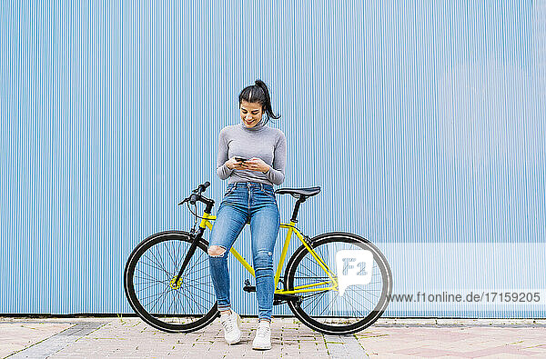 Smiling woman using mobile phone while sitting on fixie bike against blue wall
