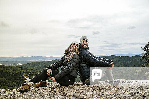 Lächelndes Paar sitzt Rücken an Rücken auf dem Aussichtspunkt gegen den Himmel