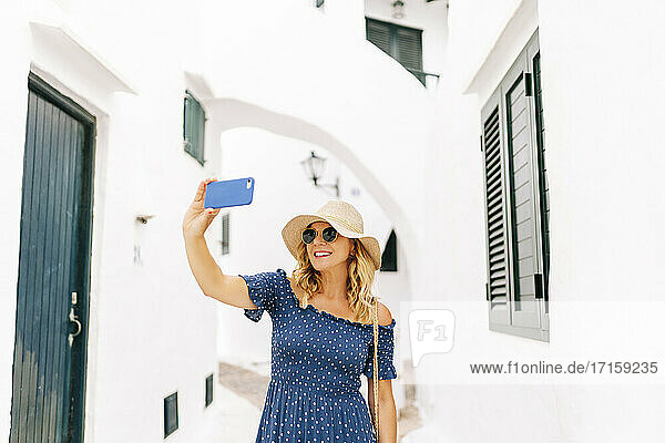 Frau mit Hut macht Selfie mit Smartphone inmitten von Häusern im Dorf Binibeca  Menorca  Spanien
