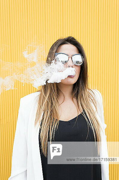 Young woman exhaling smoke against yellow wall