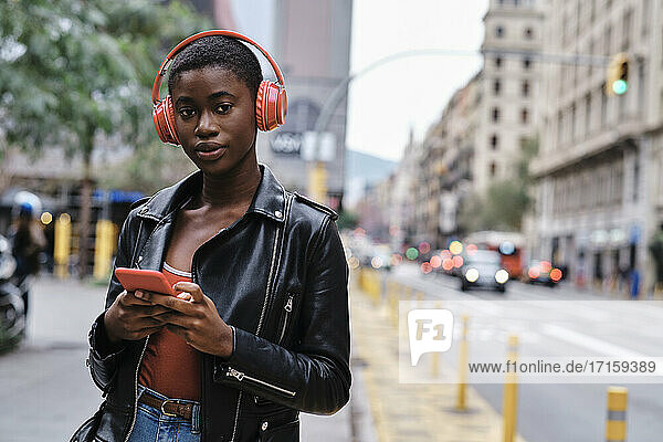 Woman wearing headphones and jacket using smart phone while standing in city