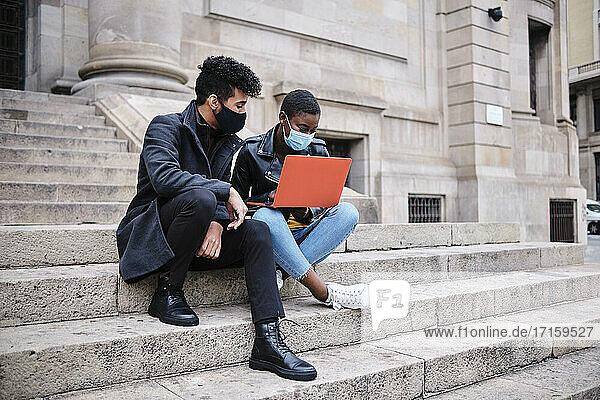 Friends wearing protective face mask using laptop while sitting on steps in city