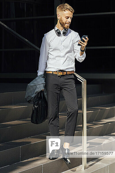 Businessman holding briefcase and jacket while using smart phone standing on steps